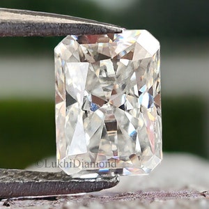 May include: A clear, rectangular, faceted gemstone with a square shape, held by tweezers. The gemstone is a radiant cut diamond.