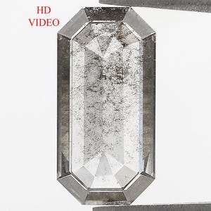 Op de afbeelding: Een emerald-cut zout en peper diamant met een rechthoekige vorm en gefacetteerd oppervlak, met een grijs en wit gespikkeld uiterlijk. De edelsteen wordt vastgehouden door een metalen pincet en bevat de rode tekst HD VIDEO linksboven.