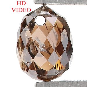 Może przedstawiać: Fasetowany, brązowy kamień szlachetny w kształcie łzy z wywierconym otworem u góry jest trzymany przez pęsetę. Kamień ma złożony, geometryczny szlif, odbijający światło. Widoczny jest tekst "HD VIDEO".