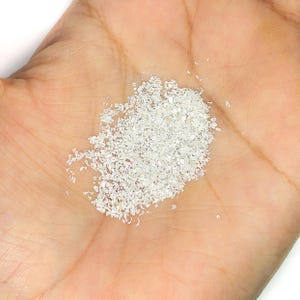 Pu&ograve; includere: Un piccolo mucchio di fini particelle di glitter bianco iridescente con una consistenza cristallina riflettente. I singoli pezzi sono di forma e dimensioni irregolari, creando un effetto scintillante quando la luce colpisce la superficie.