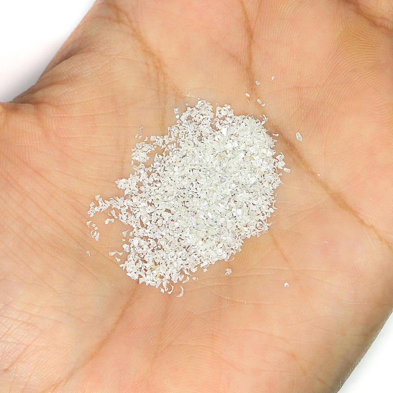 Pu&ograve; includere: Un piccolo mucchio di fini particelle di glitter bianco iridescente con una consistenza cristallina riflettente. I singoli pezzi sono di forma e dimensioni irregolari, creando un effetto scintillante quando la luce colpisce la superficie.