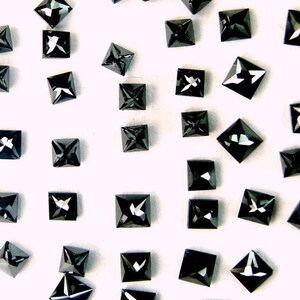 Puede incluir: Una colección de piedras preciosas negras de forma cuadrada con un patrón de facetas en forma de estrella. Las piedras preciosas están dispersas sobre una superficie blanca.
