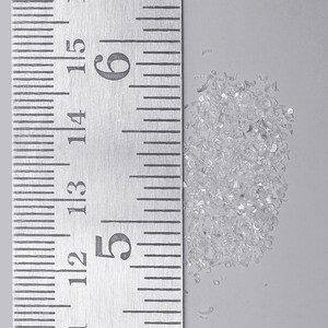 Pu&ograve; includere: Un piccolo mucchio di minuscole schegge di vetro trasparente o cristalli frantumati posizionato accanto a un righello di metallo. Il righello mostra segni in centimetri, con il mucchio che occupa circa 1,3 centimetri di altezza sulla scala da 12 a 15.