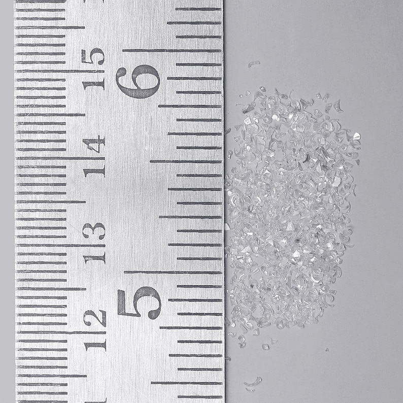 Pu&ograve; includere: Un piccolo mucchio di minuscole schegge di vetro trasparente o cristalli frantumati posizionato accanto a un righello di metallo. Il righello mostra segni in centimetri, con il mucchio che occupa circa 1,3 centimetri di altezza sulla scala da 12 a 15.
