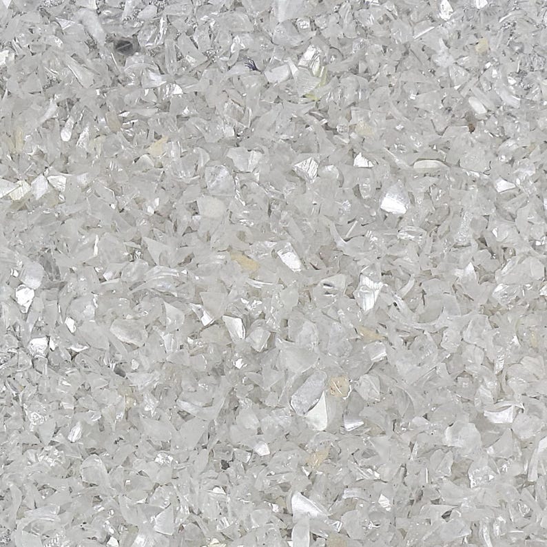Pu&ograve; includere: Una collezione di piccoli frammenti di vetro bianco traslucido e irregolare con una superficie riflettente e scintillante. I pezzi variano per dimensioni e forma, creando uno strato denso e strutturato di materiale frantumato che cattura la luce.