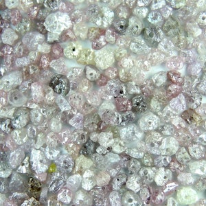 May include: A close-up view of a collection of rough, unpolished diamonds. The diamonds are various shades of white, pink, and gray. They are small and have a variety of shapes and sizes.