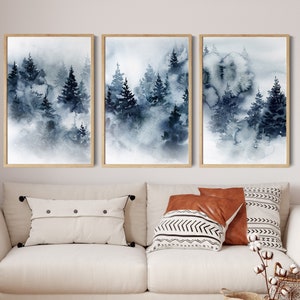 May include: Three framed watercolor paintings depict a misty forest scene in shades of blue and gray. The artwork features tall evergreen trees and a soft, atmospheric effect. The frames are a light wood color.