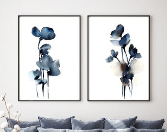 May include: Two framed watercolor floral prints in black frames. The artwork features abstract blue flowers on a white background. The prints are displayed above a blue velvet sofa with matching pillows, a round ottoman, and a floor lamp.
