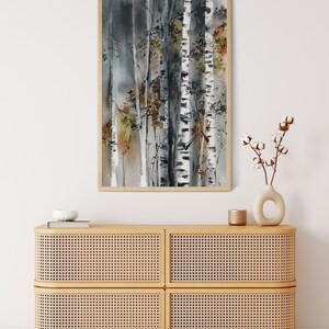 May include: Watercolor painting of birch trees in a wooden frame. The artwork features a muted color palette of gray, white, and brown, with touches of green and orange. The painting is displayed above a light wood cabinet with woven panels.