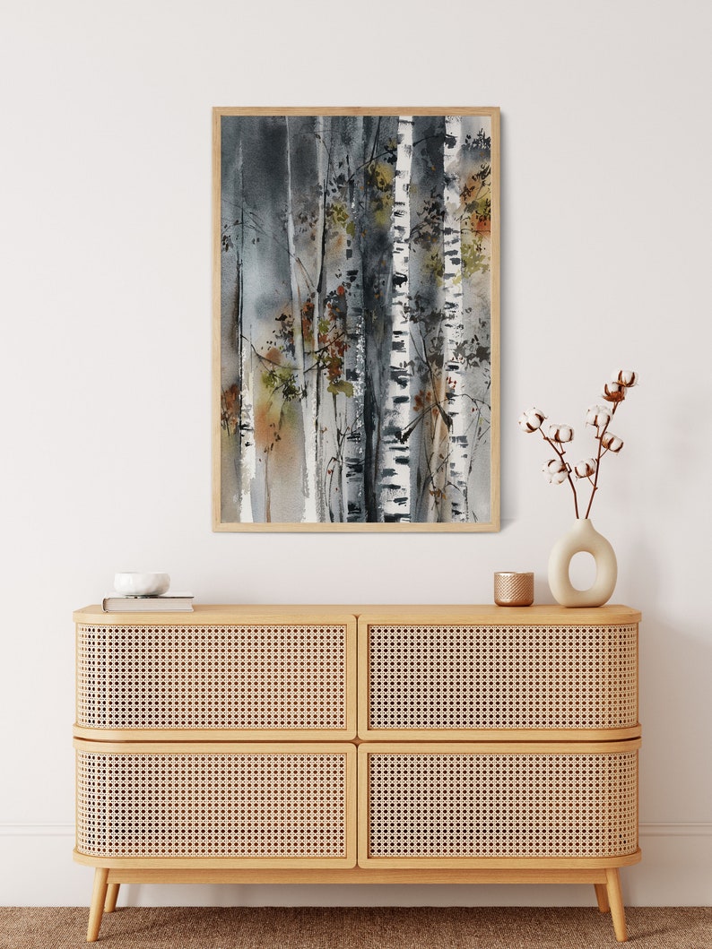 May include: Watercolor painting of birch trees in a wooden frame. The artwork features a muted color palette of gray, white, and brown, with touches of green and orange. The painting is displayed above a light wood cabinet with woven panels.