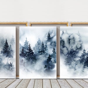 May include: Three framed watercolor paintings depict a misty forest scene. The artwork features dark blue and gray evergreen trees in a foggy landscape. The frames are a light wood color.