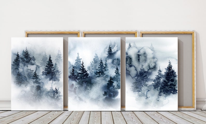 May include: Three framed watercolor paintings depict a misty forest scene. The artwork features dark blue and gray evergreen trees in a foggy landscape. The frames are a light wood color.