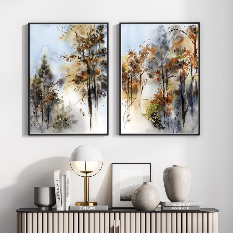 Pu&ograve; includere: Due dipinti ad acquerello incorniciati di una scena di foresta con alberi nei toni del marrone, verde e oro su uno sfondo azzurro e grigio. I dipinti sono esposti sopra una moderna consolle con oggetti decorativi.
