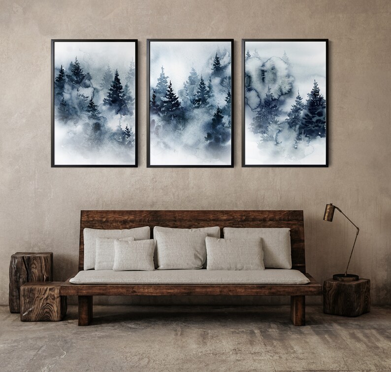 May include: Three framed watercolor prints depicting a misty forest scene in shades of blue and gray. The artwork is displayed above a wooden bench with gray cushions, complemented by a wooden side table and a modern lamp.