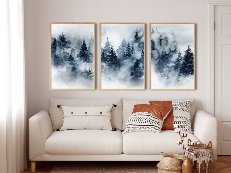 May include: Three framed watercolor paintings depict a misty forest scene in shades of blue and gray. The artwork features tall evergreen trees and a soft, atmospheric effect. The frames are a light wood color.