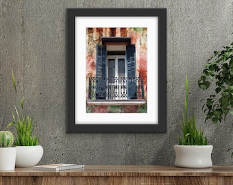 May include: Framed print of a balcony with black wrought iron railing, blue shutters, and a white door. The building's exterior has a weathered, colorful appearance. The print is displayed on a wooden surface with plants.