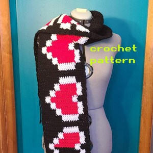 Puede incluir: Una bufanda de crochet negra, blanca y roja con flecos. La bufanda presenta un patrón repetido de corazones. El texto "crochet pattern" es visible en la imagen.