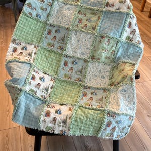 May include: A rag quilt featuring a patchwork design of light blue, sage green, and white fabric squares. The quilt displays a repeating pattern of Peter Rabbit characters, plaid textures, and floral motifs with frayed, exposed seams between squares.