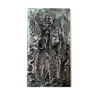 May include: A rectangular metal relief sculpture depicting a man in a detailed, ornate outfit. The figure holds a feather and a small knife. The color is a muted green with darker shadows, creating a textured appearance. The man has a flower on his jacket.