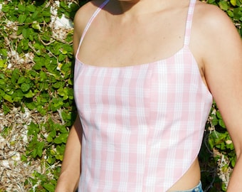May include: A sleeveless handkerchief-style top featuring a light pink and white plaid pattern. The garment has a square neckline, thin spaghetti straps, and a pointed hemline that tapers to a center point at the waist, paired with blue denim jeans.