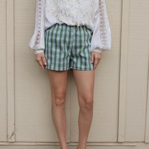 May include: A white long-sleeved button-down shirt featuring intricate lace panels on the chest and sleeves, paired with green and white plaid shorts. The shirt has a collar and full-length sleeves, while the shorts feature a classic checkered pattern.