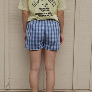 May include: A pale yellow short-sleeve t-shirt with black text reading THE HANGER CLUB & RECREATION CENTER PALMEROLA AIR HONDURAS, C. and blue and white plaid shorts paired with white socks and pink sneakers with blue stripes and gum soles.
