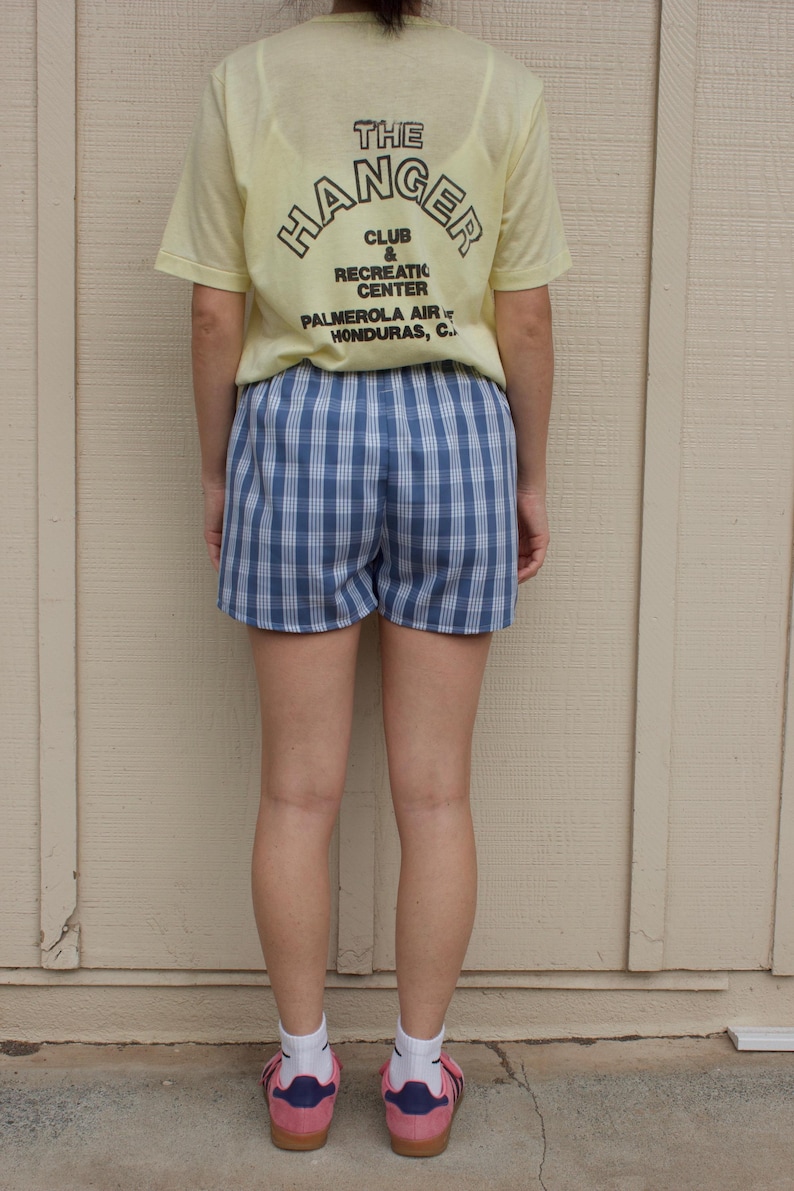 May include: A pale yellow short-sleeve t-shirt with black text reading THE HANGER CLUB & RECREATION CENTER PALMEROLA AIR HONDURAS, C. and blue and white plaid shorts paired with white socks and pink sneakers with blue stripes and gum soles.