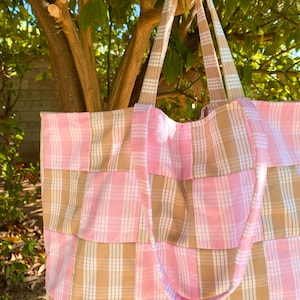 May include: A large tote bag with a pink and tan plaid pattern. The bag has long straps and is made of fabric. The bag is hanging from a tree branch, with green leaves in the background.