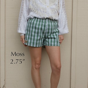 May include: Green and white plaid shorts featuring a small embroidered hand gesture patch on the lower right leg. The shorts are labeled Moss and include a 2.75 inch measurement text. The garment has an elastic waistband and a straight hemline.