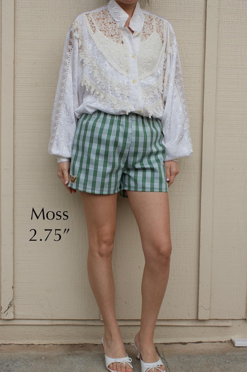 May include: Green and white plaid shorts featuring a small embroidered hand gesture patch on the lower right leg. The shorts are labeled Moss and include a 2.75 inch measurement text. The garment has an elastic waistband and a straight hemline.