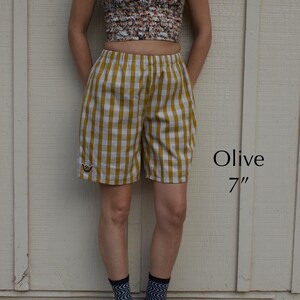 May include: Olive and white gingham shorts with an elastic waistband and a small embroidered hand gesture patch on the lower left leg. The text Olive 7" is visible to the right of the shorts, which feature a classic checkered pattern.