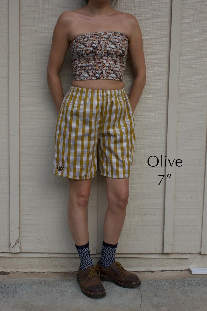 May include: Olive and white gingham shorts with an elastic waistband and a small embroidered hand gesture patch on the lower left leg. The text Olive 7" is visible to the right of the shorts, which feature a classic checkered pattern.