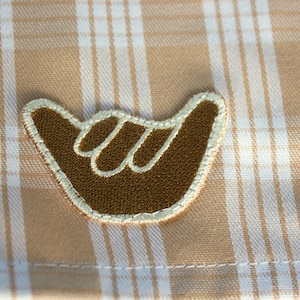 May include: A brown shaka hand sign embroidered patch with a cream-colored stitched border, positioned on a tan and white plaid fabric background. The patch features a textured surface and a distinct outline of the fingers in a lighter thread.