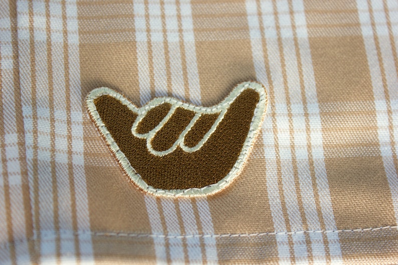May include: A brown shaka hand sign embroidered patch with a cream-colored stitched border, positioned on a tan and white plaid fabric background. The patch features a textured surface and a distinct outline of the fingers in a lighter thread.