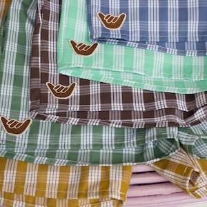 May include: A collection of plaid shorts in mustard yellow, sage green, dark brown, and light blue. Each pair of shorts features a white and brown shaka hand sign patch sewn onto the lower corner. The fabric displays a consistent grid pattern.