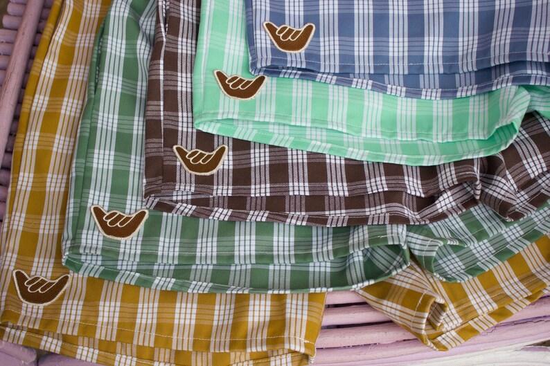 May include: A collection of plaid shorts in mustard yellow, sage green, dark brown, and light blue. Each pair of shorts features a white and brown shaka hand sign patch sewn onto the lower corner. The fabric displays a consistent grid pattern.