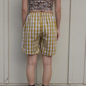 May include: Mustard yellow and white gingham shorts featuring an elastic waistband. The shorts have a relaxed fit and reach mid-thigh. The fabric pattern consists of a consistent grid of vertical and horizontal lines in a classic checkered style.