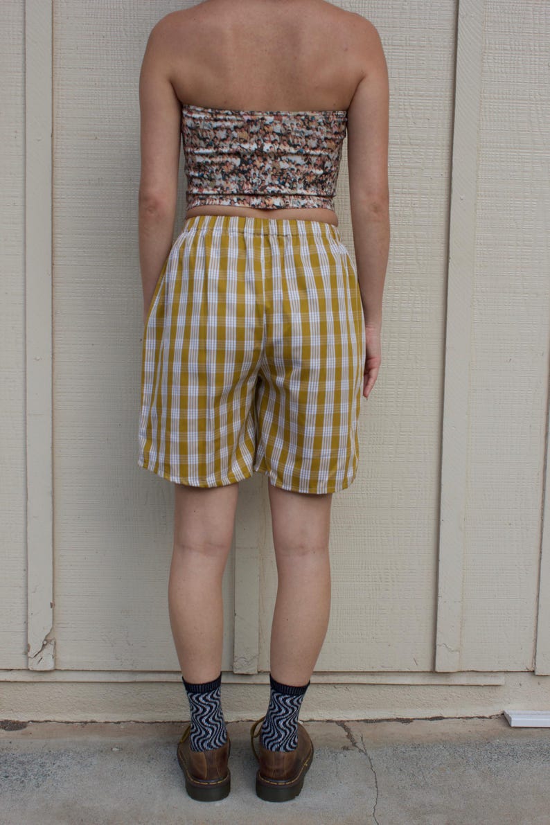 May include: Mustard yellow and white gingham shorts featuring an elastic waistband. The shorts have a relaxed fit and reach mid-thigh. The fabric pattern consists of a consistent grid of vertical and horizontal lines in a classic checkered style.
