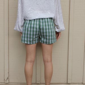 May include: A white long-sleeve blouse with a subtle tonal floral pattern paired with green and white plaid shorts. The blouse features loose sleeves and a relaxed fit, while the shorts have a classic checkered design and a simple waistband.