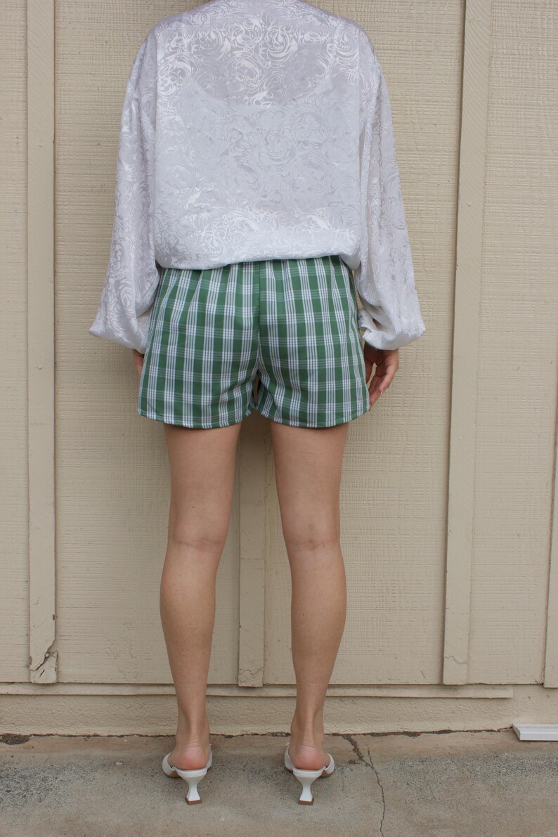 May include: A white long-sleeve blouse with a subtle tonal floral pattern paired with green and white plaid shorts. The blouse features loose sleeves and a relaxed fit, while the shorts have a classic checkered design and a simple waistband.