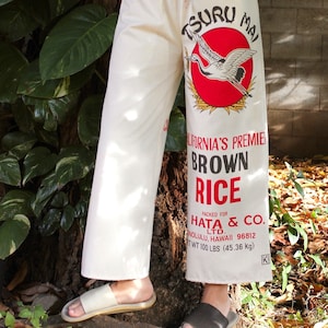 May include: White pants with a red and white graphic of a crane and the text "Tsuru Mai California's Premier Brown Rice Packed for Hata & Co. Ltd. Honolulu, Hawaii 96812 Net Wt 100 Lbs (45.36 kg)"
