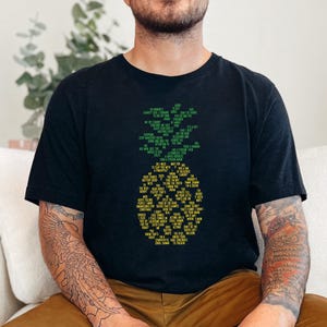May include: A black short-sleeved t-shirt featuring a graphic of a pineapple composed of small green and yellow text. The pineapple shape is centered on the chest, with the green text forming the leaves and the yellow text forming the fruit body.