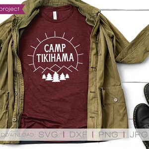 May include: A maroon t-shirt with the text "CAMP TIKIHAMA" in a white graphic design, featuring a sun, mountains, and trees. The shirt is displayed with a green jacket, black shoes, and a small plant on a white wooden surface.