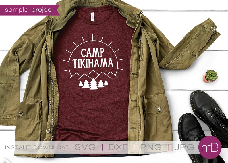 May include: A maroon t-shirt with the text "CAMP TIKIHAMA" in a white graphic design, featuring a sun, mountains, and trees. The shirt is displayed with a green jacket, black shoes, and a small plant on a white wooden surface.