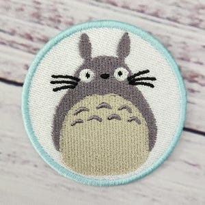 May include: Embroidered patch featuring a cartoon Totoro character. The patch is round, with a light blue border and a white background. The Totoro is gray with a beige belly, black whiskers, and large eyes. A cute, whimsical design.