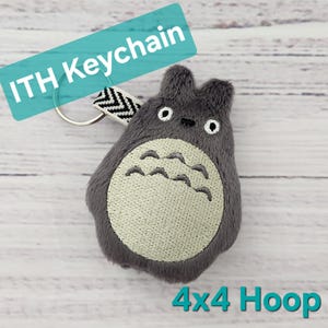 Może przedstawiać: Szary pluszowy brelok do kluczy w kształcie postaci z kreskówki, z białym haftowanym brzuchem i rysami twarzy. Posiada srebrny pierścień i czarno-biały pasek. Widoczny jest tekst "ITH Keychain" i "4x4 Hoop".