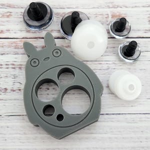 May include: A gray, Totoro-shaped craft tool with multiple circular openings, alongside several black-and-clear plastic eye components and white plastic doll parts. The items are arranged on a light-colored wooden surface.