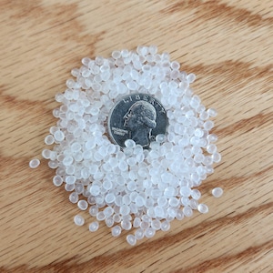 May include: A collection of small, translucent, circular plastic pellets scattered in a pile. A silver-colored United States quarter coin is placed in the center of the pellets to provide a sense of scale for the size of the individual plastic beads.
