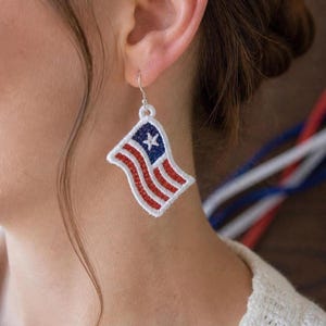 以下が含まれることがあります： アメリカ国旗のデザインが施された刺繍の旗のイヤリング。イヤリングは白い縁取り、赤と白のストライプ、白い星が付いた青い角があります。銀色のフックから吊り下げられています。