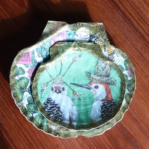 Puede incluir: Un cuenco decorativo en forma de concha. El cuenco presenta una imagen central de dos pájaros con coronas sobre un fondo verde. Los bordes están adornados con patrones dorados y florales. El cuenco está sobre una superficie de madera.